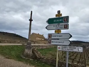 Directional signpost with multiple arrows pointing toward various French towns, set against a backdrop of a rural landscape with cloudy skies, leading to locations for a Burgundy wine tasting tour.