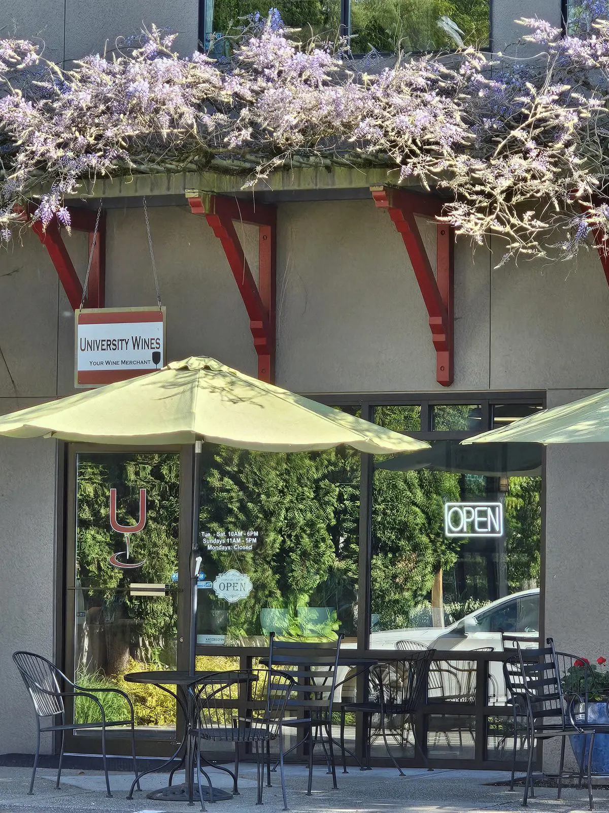 Outdoor seating area of University Wines with an “open” sign, patio umbrella, chairs, and blooming purple tree above.