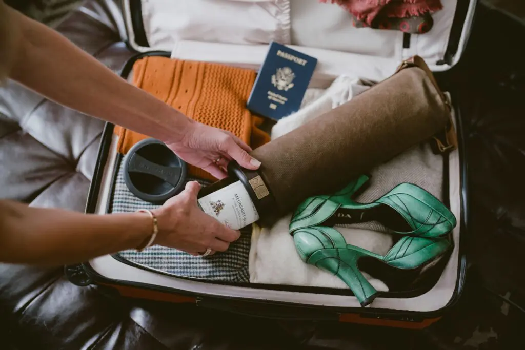 A Vinarmour wine carrier is placed inside of a neatly packed suitcase filled with clothes and a passport. Someone is placing or removing a bottle of wine from the carrier.