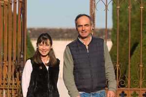 A couple smiling at the camera, standing behind an ornate metal gate in a sunny outdoor setting at Château Couronneau.