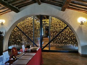 View through an arched doorway of a Tuscan wine cellar with numerous hanging wine glasses, table with wine bottles, and warm ambient lighting.