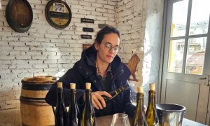 Woman in glasses and coat sitting at a table with Burgundy wine bottles, examining a wine glass in a rustic room with white brick walls.