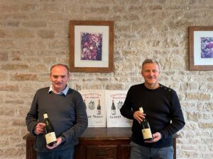 Two men stand in front of a light cream colored brick/stone wall. They each hold a bottle of wine. They stand in front of a wooden cabinet and framed photos adorn the walls.