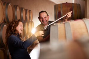 Woman and man among barrels of wine in a cellar tasting one of the wines.