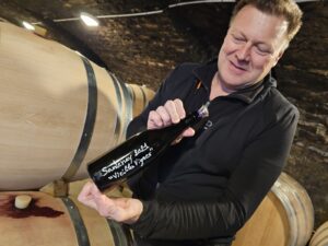 A man displays a 2021 bottle of Domaine Gabriel & Paul Jouard wine to the camera in a cellar.