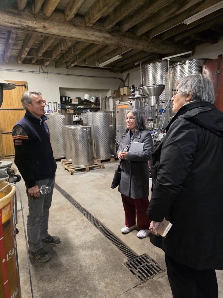 Three people standing and discussing wine with wine production equipment in the background.