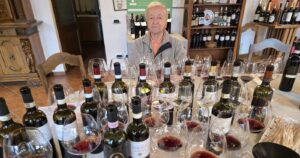 A man sits behind a large assortment of wine bottles and glasses sprawled all over a table.