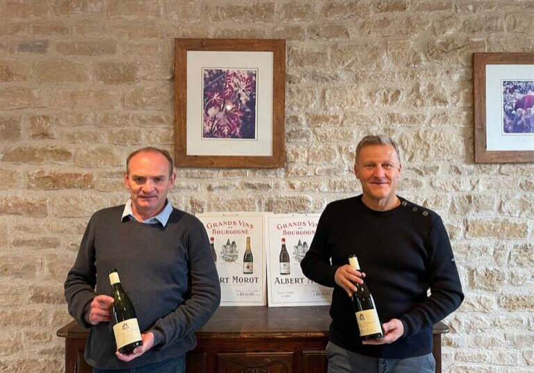 Two men stand in front of a light cream colored brick/stone wall. They each hold a bottle of wine. They stand in front of a wooden cabinet and framed photos adorn the walls.