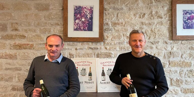 Two men stand in front of a light cream colored brick/stone wall. They each hold a bottle of wine. They stand in front of a wooden cabinet and framed photos adorn the walls.