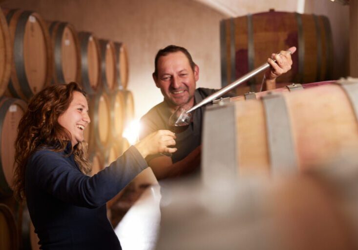 Woman and man among barrels of wine in a cellar tasting one of the wines.