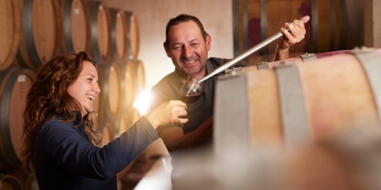 Woman and man among barrels of wine in a cellar tasting one of the wines.