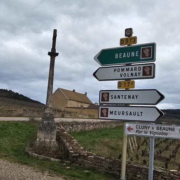 Directional signpost with multiple arrows pointing toward various French towns, set against a backdrop of a rural landscape with cloudy skies, leading to locations for a Burgundy wine tasting tour.