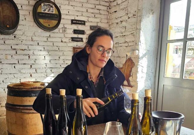 Woman in glasses and coat sitting at a table with Burgundy wine bottles, examining a wine glass in a rustic room with white brick walls.