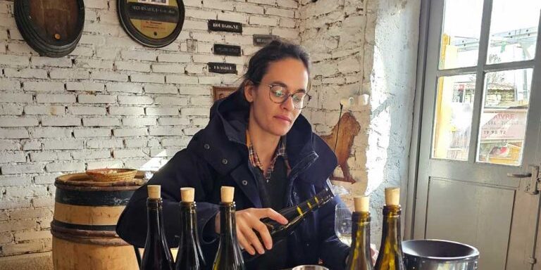 Woman in glasses and coat sitting at a table with Burgundy wine bottles, examining a wine glass in a rustic room with white brick walls.