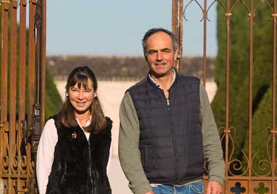 A couple smiling at the camera, standing behind an ornate metal gate in a sunny outdoor setting at Château Couronneau.