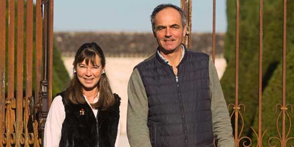 A couple smiling at the camera, standing behind an ornate metal gate in a sunny outdoor setting at Château Couronneau.