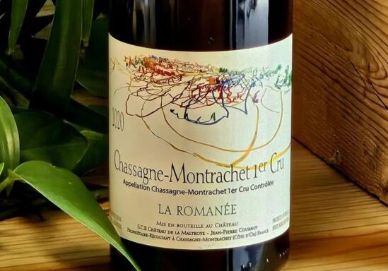 A bottle of wine resting against a wooden background with vibrant green houseplant nestled nearby. This image shows the bottle of Château de la Maltroye 2020 Chassagne-Montrachet 1er Cru La Romanée.