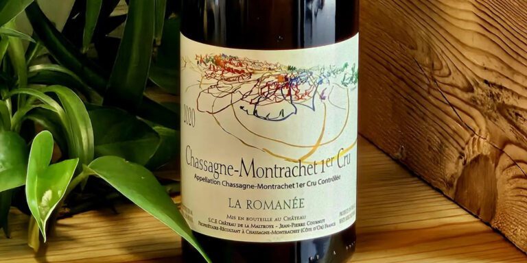 A bottle of wine resting against a wooden background with vibrant green houseplant nestled nearby. This image shows the bottle of Château de la Maltroye 2020 Chassagne-Montrachet 1er Cru La Romanée.