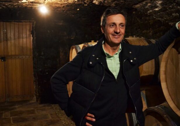 A man stands in a cellar at Domaine Buisson Battault with his hand resting on a barrel.