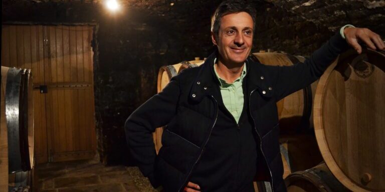 A man stands in a cellar at Domaine Buisson Battault with his hand resting on a barrel.