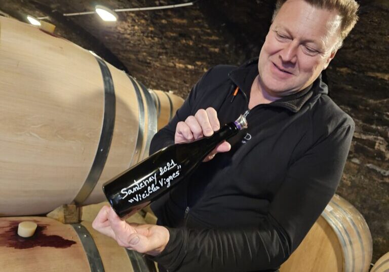 A man displays a 2021 bottle of Domaine Gabriel & Paul Jouard wine to the camera in a cellar.