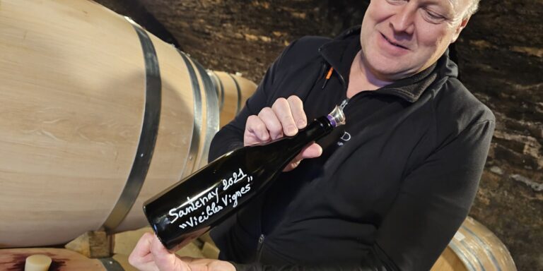 A man displays a 2021 bottle of Domaine Gabriel & Paul Jouard wine to the camera in a cellar.