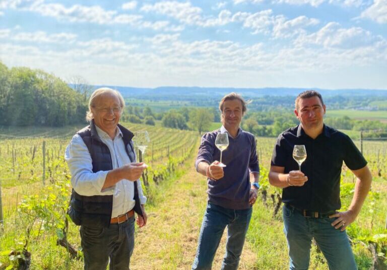 The family in charge of the Domaine de Sainte Marie raises their glasses towards the camera in a vineyard under a blue sky.