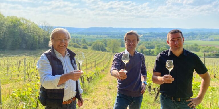 The family in charge of the Domaine de Sainte Marie raises their glasses towards the camera in a vineyard under a blue sky.