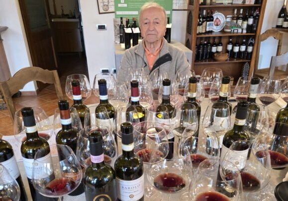 A man sits behind a large assortment of wine bottles and glasses sprawled all over a table.