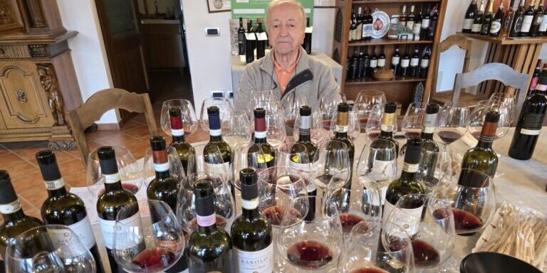 A man sits behind a large assortment of wine bottles and glasses sprawled all over a table.