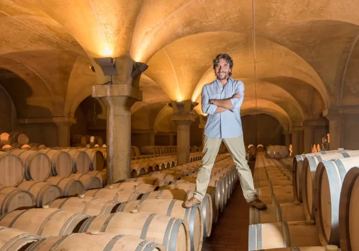 A man with a beard smiles and stands with arms crossed in a wine cellar surrounded by wooden barrels under arched ceilings.