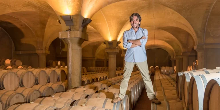 A man with a beard smiles and stands with arms crossed in a wine cellar surrounded by wooden barrels under arched ceilings.