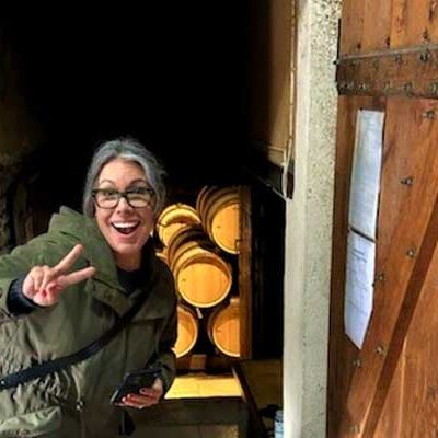 Michelle Schaber in glasses and a green jacket gives a peace sign while standing in a doorway leading to a room filled with wooden barrels at University Wines.