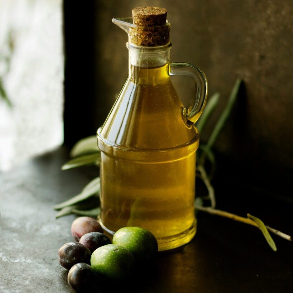 An image of olive oil on a table with olives besides it.